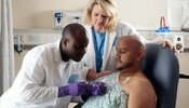 A Hispanic male patient receives Chemotherapy from a African-American Nurse through a port that is placed in his chest area. A caucasian female nurse looks on.